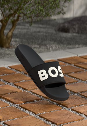 Sort slidesandal med hvidt "BOSS" bogstaver, der svæver over trækvadrattfliser med små sten imellem.