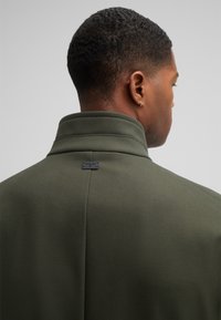 Olijfgroen jasje met hoge kraag, textuurstof en een klein zwart label op de achterkant. Glad, op maat gemaakt ontwerp met subtiele lijnen.