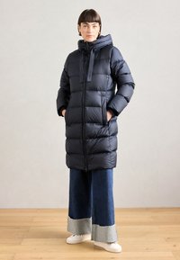 Puff jakke i marineblått med hette, med glidelås foran, quiltet design og to sidelommer. Kombinert med vide denimbukser.