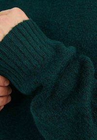 Teal stickad tröja med detaljer, ribbade ärmslut och mjuk textur. Ärmsluten är något lösa, vilket visar en hand som greppar tyget.