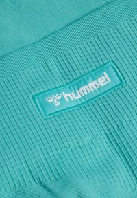 Turkis ribbet stof med et påsyet rektangulært logo, der viser en stiliseret bi og mærkenavnet "hummel" i hvid.