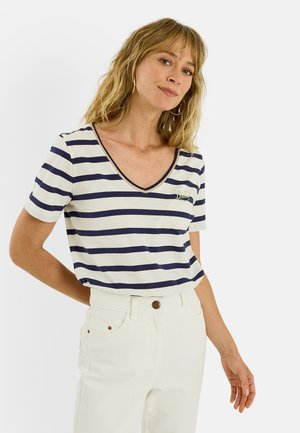 Mujer con cabello rubio que lleva una camiseta blanca con rayas azul marino con cuello en V y pantalones blancos, de pie con una mano detrás de la espalda.