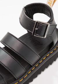 Sandale en cuir noir avec deux larges brides et bride de cheville dotée d'une boucle argentée embossée du texte "Dr. Martens", sur une semelle noire avec coutures jaunes.