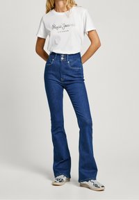 Camiseta blanca con logo impreso, jeans acampanados de cintura alta en azul, diseño de cinco bolsillos, zapatillas plateadas con acentos brillantes.