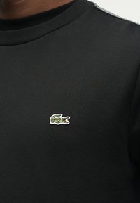 Κοντινή λήψη μαύρου πουκάμισου με μικρό κεντητό λογότυπο κροκοδείλου σε πράσινο και λευκό στο στήθος, με κόκκινη λεπτομέρεια στο εσωτερικό του στόματος.