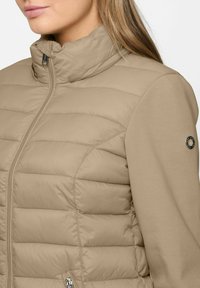 Beige gesteppte Jacke mit glatten Stoffärmeln, durchgehendem Reißverschluss, hohem Kragen und rundem Metall-Logo-Emblem am oberen Ärmel.