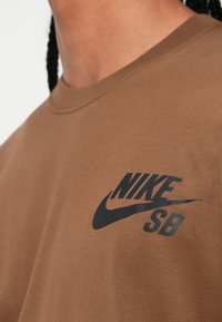 Rudas medvilninis marškinėliai su juodu Nike SB logotipu kairėje krūtinės pusėje. Paprastas apvalus iškirpimas ir švelni, tekstūruota medžiaga.