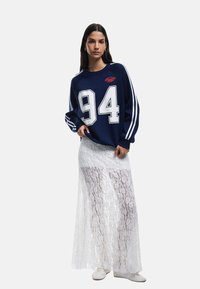 Sudadera navy con el número blanco "94" y mangas a rayas, combinada con una falda maxi de encaje blanco transparente. Modelo llevando calzado blanco.