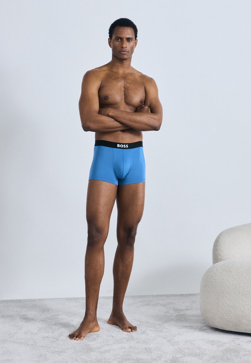 Boxers bleus avec une ceinture noire marquée "BOSS", mannequin debout les bras croisés, torse nu, fond uni.
