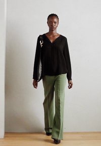 Schwarze Bluse mit langen Ärmeln und V-Ausschnitt, kombiniert mit grünen weit geschnittenen Hosen und schwarzen High Heels. Eine schwarze Tasche hängt über einer Schulter.