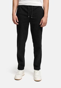 Mies, joka on pukeutunut mustiin nippusateenpohjaisiin housuihin ja valkoisiin lenkkareihin, seisoo kädet rentoina sivuillaan valkoista taustaa vasten.