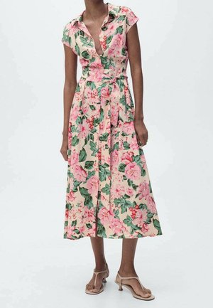 Mujer con un vestido floral sin mangas abotonado con flores rosas y hojas verdes, combinado con sandalias de tacón beige con punta abierta.