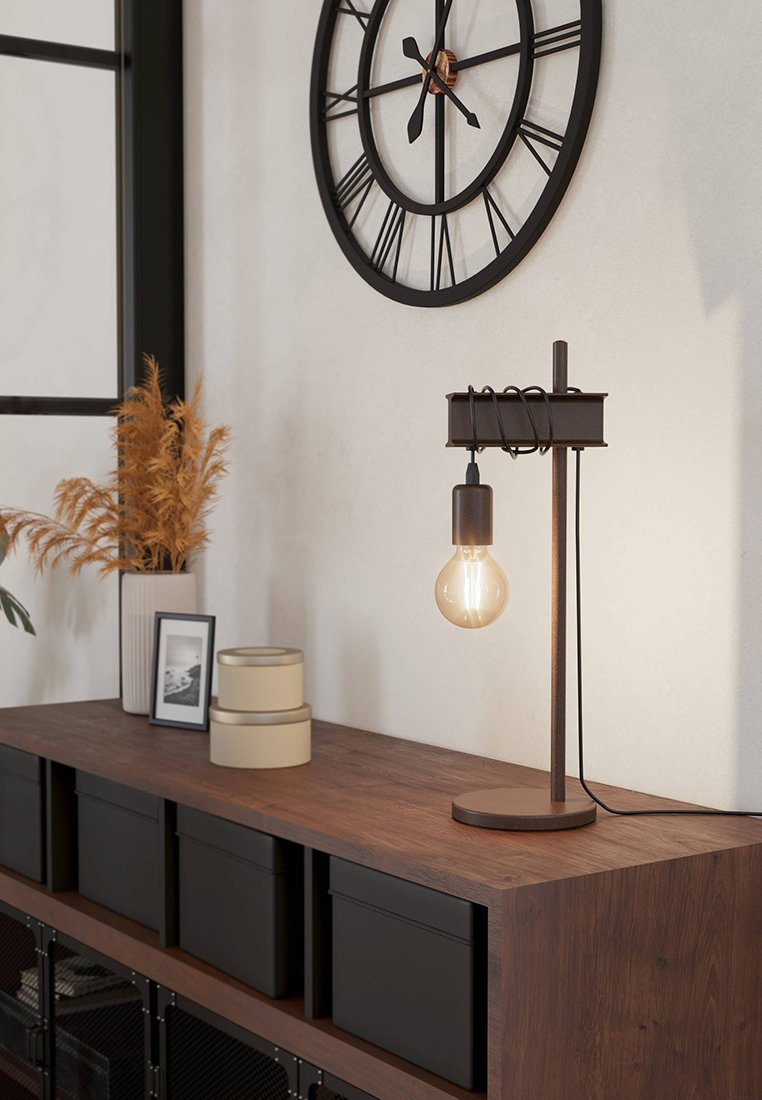 Tischlampe mit einem Holzfuß und metallischen Akzenten, ausgestattet mit einer freiliegenden Glühbirne. Umgebende Elemente sind eine Uhr, Aufbewahrungsboxen und Pflanzen.