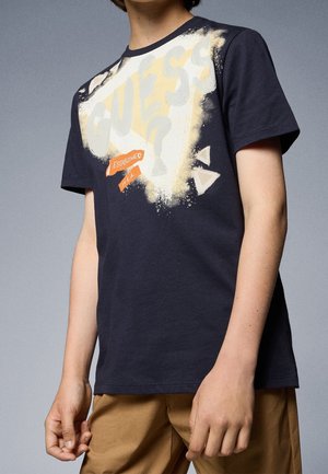 Osoba ubrana w granatowy T-shirt z dużym logo "GUESS?" i napisem "Established in L.A.", w połączeniu z jasnobrązowymi spodniami.