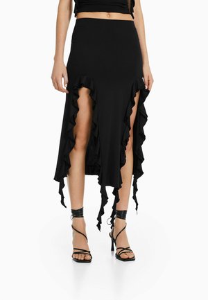 Jupe asymétrique noire avec de hautes fentes devant et un bord volanté, portée avec des sandales noires à talons hauts et à brides.