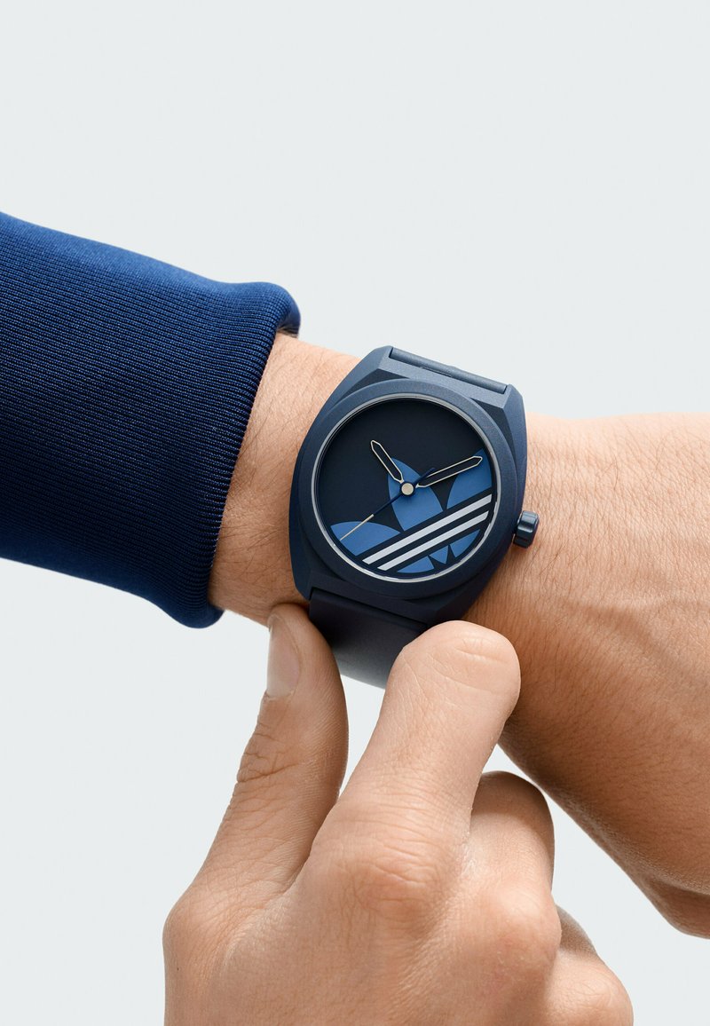 Personne ajustant une montre-bracelet bleue avec le logo Adidas sur le cadran, portant une chemise à manches longues bleu marine sur un fond uni.
