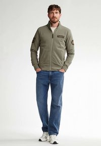 Giacca verde oliva con zip, collo alto e toppe sulle spalle, abbinata a jeans blu e sneakers bianche. Design semplice e vestibilità comoda.