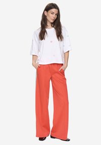 Bílý top s krátkými rukávy a barevným výšivkovým srdcem. Volnější střih, kombinovaný s oranžovými kalhotami s širokými nohavicemi a vysokým pasem, zdobenými bočními pruhy.