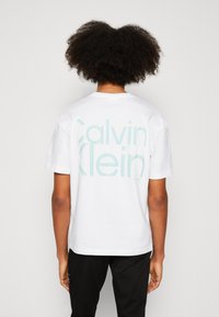 Bílá bavlněná tričko s velkým mátově zeleným logem "Calvin Klein" potištěným na zadní straně. Design s kulatým výstřihem a krátkými rukávy.