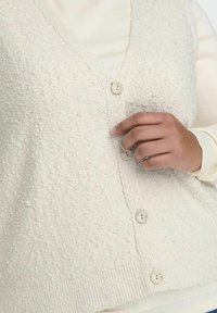 Krämfärgad, texturerad stickad väst med djup V-ringning, fem knappar och ribbad fåll. Närbild som framhäver tygets taktila kvalitet.