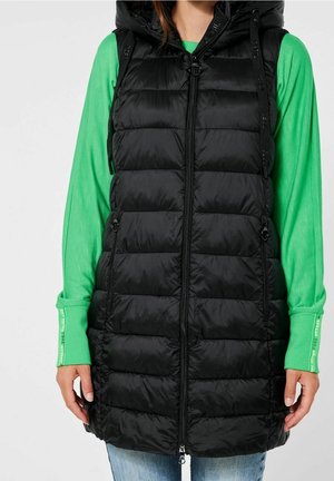Gilet rembourré noir avec capuche, présentant une fermeture éclair sur le devant, des poches latérales et une texture de tissu lisse et brillant. Porté par-dessus une chemise à manches longues verte.