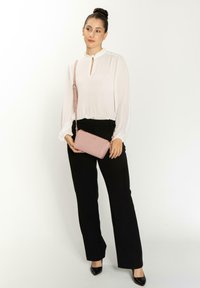 Borsa a tracolla rosa chiaro con finitura texturizzata, hardware dorato e tracolla rimovibile. Il modello indossa pantaloni neri e una blusa bianca.