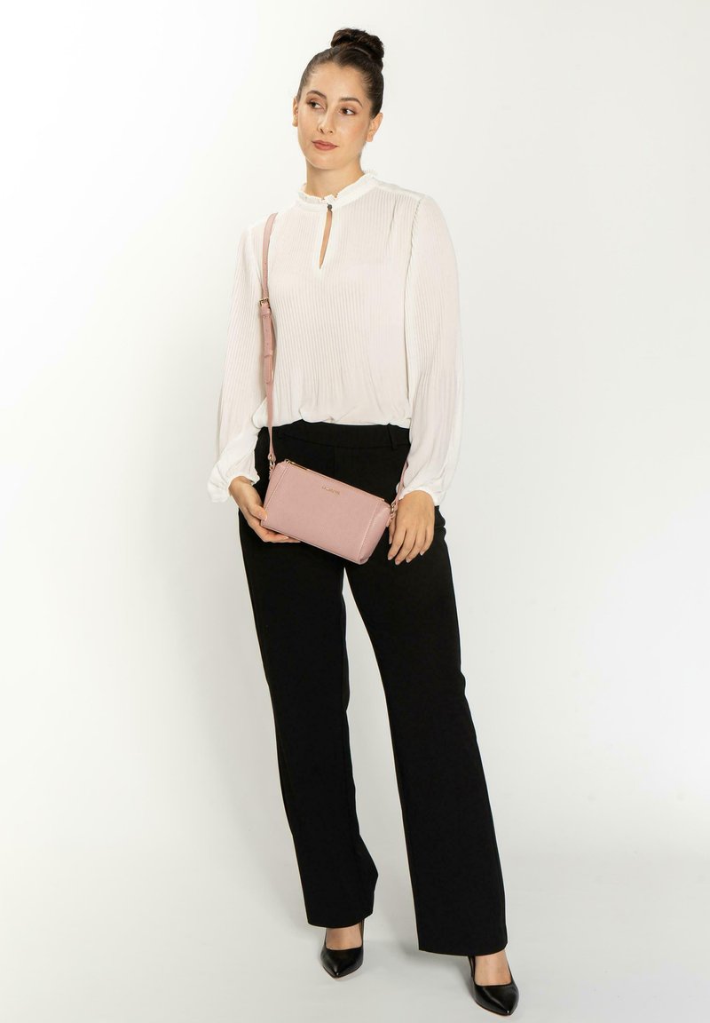Borsa a tracolla rosa chiaro con finitura texturizzata, hardware dorato e tracolla rimovibile. Il modello indossa pantaloni neri e una blusa bianca.
