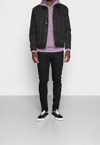 Svart denimjacka med två framfickor som överlappar en ljuslila hoodie, kombinerad med svarta jeans och svarta sneakers med vita sulor.