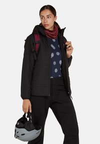 Svart pufferjacka med quiltat mönster, bärs över en marinblå tröja med ljusa prickar. Har även mörka byxor och en grå hjälm.