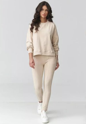 Kobieta z długimi kręconymi włosami, ubrana w beżową bluzę i spodnie, idzie do przodu na jasnym, jednolitym tle. Widoczne białe trampki.