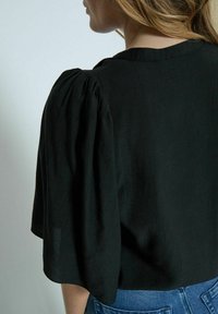 Crna bluzica s napuhanim kratkim rukavima, napravljena od mekog materijala. Ima okrugli izrez i slobodan kroj, uparen s plavim trapericama.