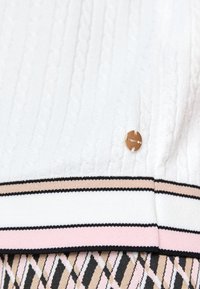 Tissu en maille torsadée blanche avec un emblème rond doré, bordé de rayures roses et beiges au-dessus d’un motif géométrique beige, noir et rose.