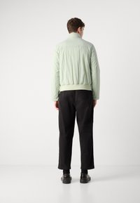 Jachetă bomber verde deschis cu un finisaj texturat, cu manșete și tiv ribbate, asortată cu pantaloni negri și încălțăminte neagră.