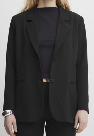 Frau trägt einen schwarzen Blazer und ein geripptes schwarzes Crop-Top, zeigt den Bauch und ein silbernes Armband am linken Handgelenk vor einfarbigem Hintergrund.