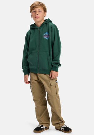 Groene rits hoodie met voorzakken, voorzien van een multiculturele grafische print. Gecombineerd met beige cargobroek en zwarte sneakers.