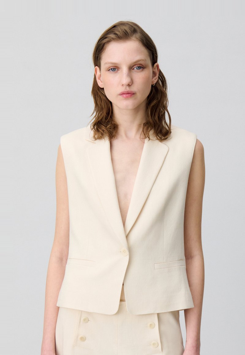 Blazer sans manches beige avec un décolleté en V profond et une fermeture à un bouton. Fabriqué en tissu texturé, il présente des poches latérales et une coupe ajustée.