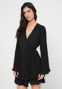 Μαύρο πλισέ φόρεμα τύπου wrap με μακριά, φαρδιά μανίκια και ζώνη στη μέση. Διαθέτει V-λαιμόκοψη και στρωμένο βολάν στο τελείωμα. Απαλή υφή υφάσματος.