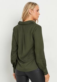 Mørkegrøn blouse med lange ærmer, krave og knapmanchetter. Har en plissering detalje på bagsiden og en glat tekstur.