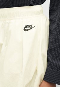 Κοντινή λήψη λευκών παντελονιών Nike με ελαστική μέση και μαύρο λογότυπο, φορεμένα με σκουρόχρωμο μακρυμάνικο πουκάμισο με υφή.