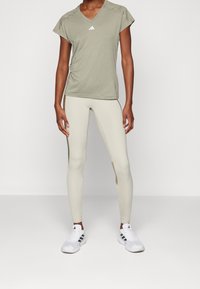 Mujer con una camiseta de cuello en V de manga corta en verde oliva claro, leggins de cintura alta en beige con paneles laterales y zapatillas blancas con franjas negras.