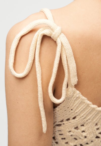 Κρεμ χρωματισμένος πλεκτός ιμάντας με κόμπους στον ώμο, προσκολλημένος σε πλεκτή μπλούζα με δαντελένιο μοτίβο.