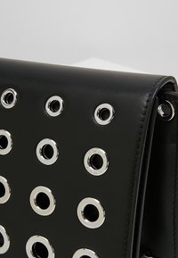 Bolso de mano de cuero negro con ojales metálicos dispuestos en un patrón de cuadrícula, con un diseño elegante y una textura suave con acentos mínimos.