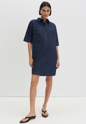 Vrouw staand, gekleed in een donkerblauwe spijkerjurk met korte mouwen, een kraag en twee borstzakken, gecombineerd met zwarte sandalen met hak.
