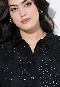 Chemise noire avec des motifs floraux brodés et des accents blancs. Elle possède un col, une fermeture à boutons et un tissu texturé.