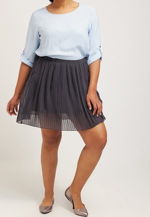 Femme portant un chemisier bleu clair à manches longues, une jupe plissée noire et des chaussures plates métalliques, se tenant devant un fond uni.