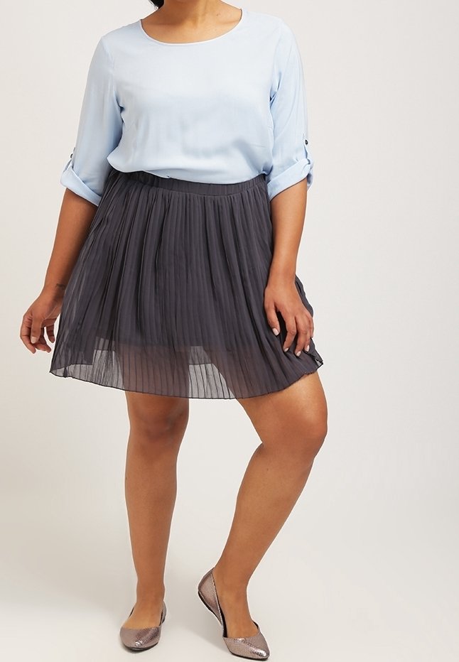 Femme portant un chemisier bleu clair à manches longues, une jupe plissée noire et des chaussures plates métalliques, se tenant devant un fond uni.