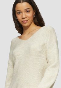 Lichtbeige gebreide trui met een V-hals en lange mouwen, voorzien van een textuurpatroon en een ontspannen pasvorm. Soepele stof, zachte uitstraling.