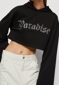 Hoodie noir court à manches longues, avec l'inscription "Paradise" en strass sur le devant. Tissu doux avec une coupe décontractée.