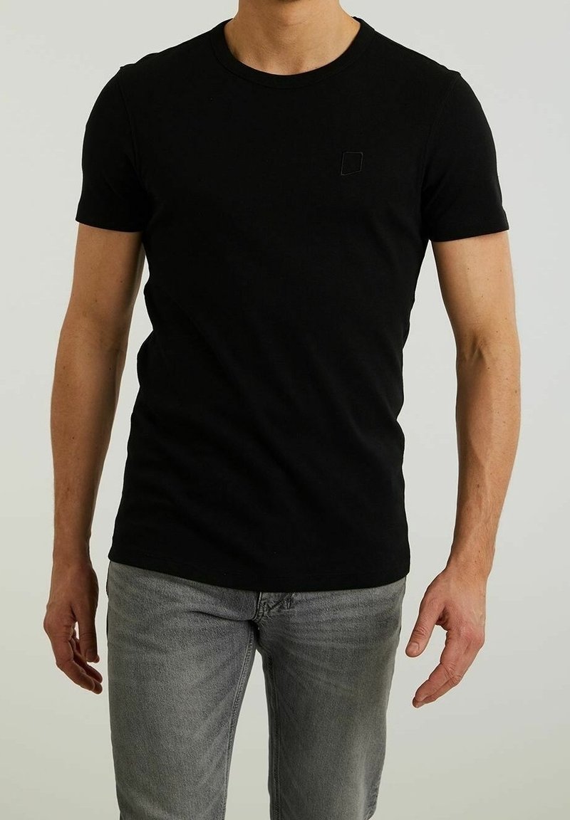 Homme portant un t-shirt noir uni à manches courtes et un jean gris clair, debout devant un fond uni.