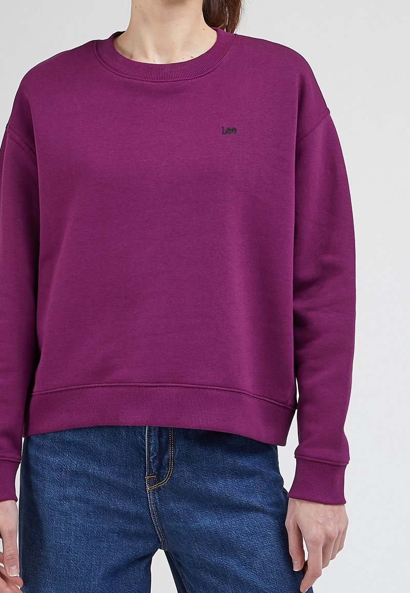 Purple sweatshirt with a round neckline and ribbed cuffs. Features a small black logo on the chest. Made from soft, textured fabric.